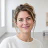 Portret van een glimlachende vrouw met opgestoken bruin haar en een witte trui in een lichte galerijzaal